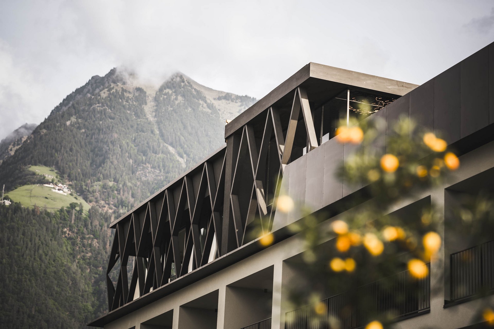 Bewusste Auszeit: Ihr Yoga-Hotel in Meran Modernes Gebäude mit Gebirgshintergrund und unscharfen orangefarbenen Blumen im Vordergrund