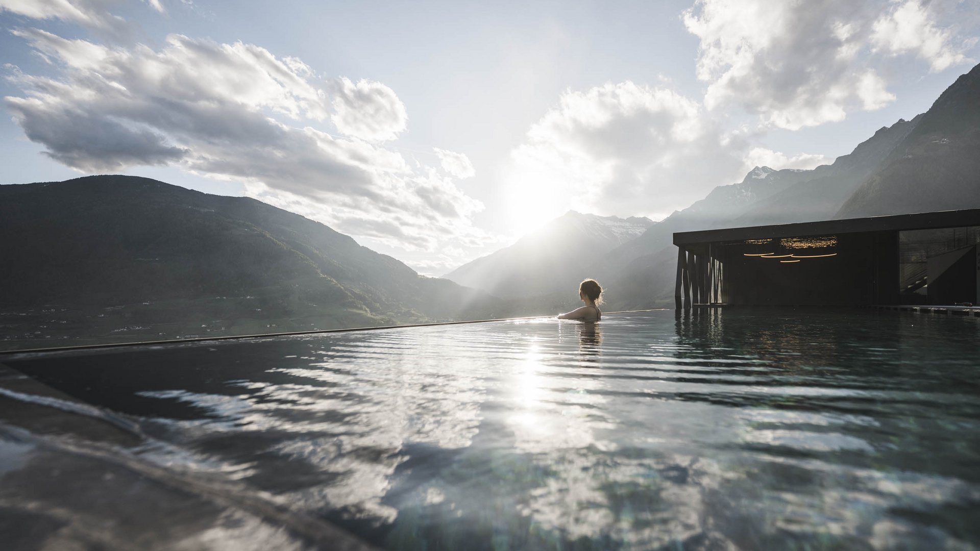 Sky Pool & Garden Pool in Dorf Tirol – Ihr Sprung ins Glück | SomVita Suites Person in Infinity-Pool mit Berglandschaft und untergehender Sonne