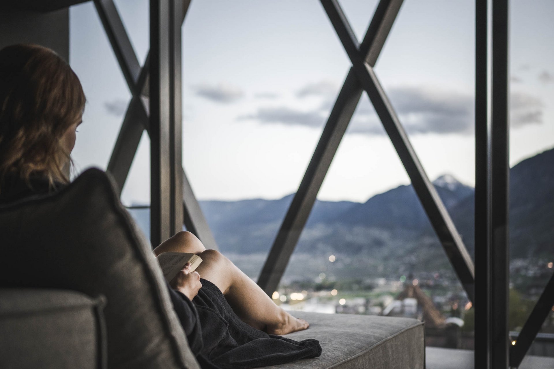 Sky Pool & Garden Pool in Dorf Tirol – Ihr Sprung ins Glück | SomVita Suites Person liest auf Sofa vor großem Fenster mit Bergblick